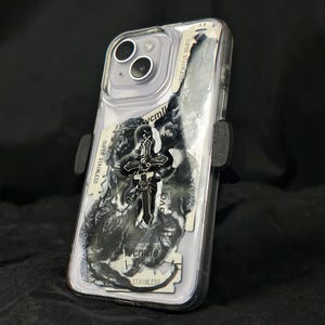 May include: Clear phone case with a black and white abstract design and a silver cross charm. The case has a black holder on the side. The words "SUPER STAINLESS" are visible on a piece of paper inside the case.