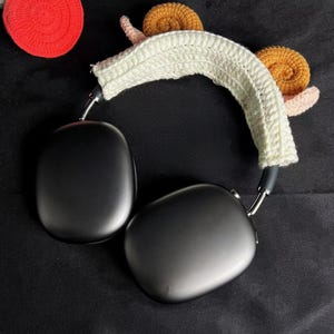 May include: Black over-ear headphones with a white crocheted headband featuring two snail designs. The snails have brown shells and pink bodies. Additional crocheted elements, including a red apple and a green flower, are visible.