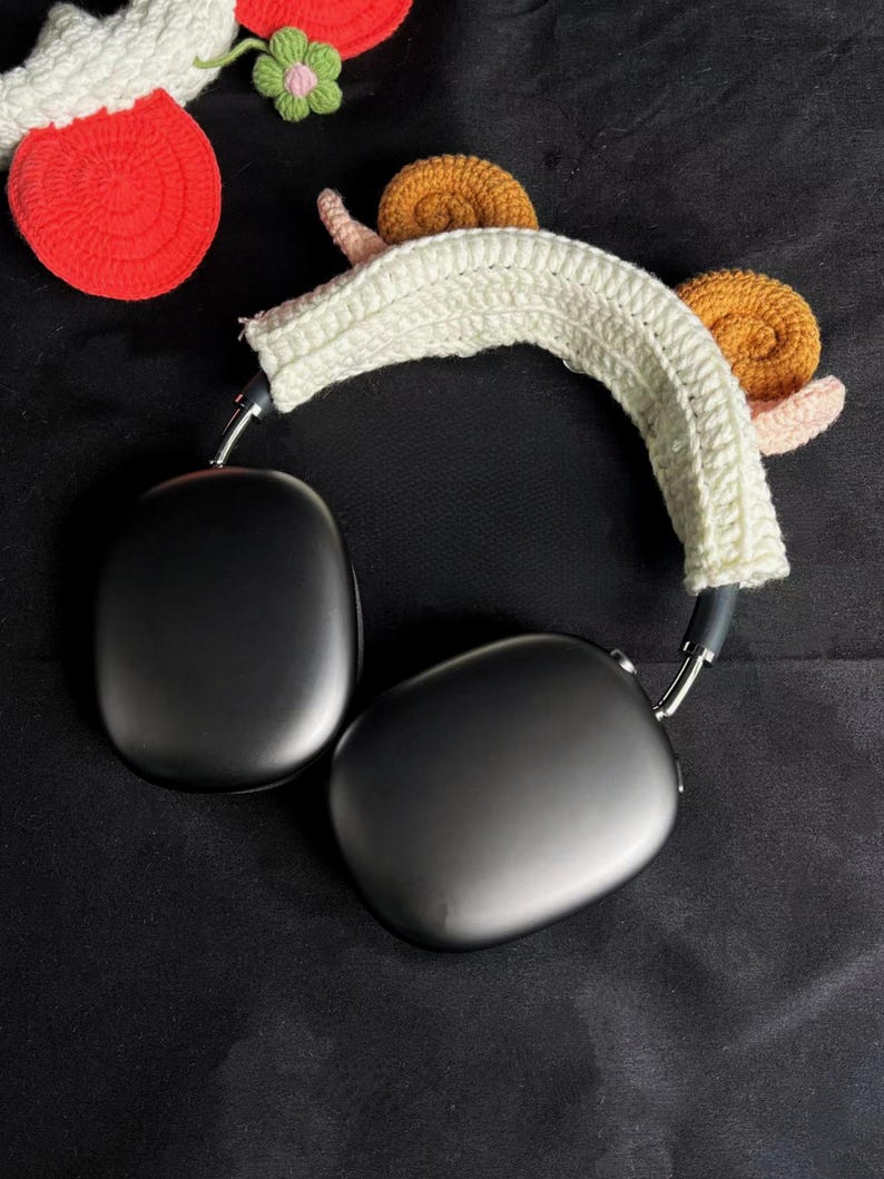 May include: Black over-ear headphones with a white crocheted headband featuring two snail designs. The snails have brown shells and pink bodies. Additional crocheted elements, including a red apple and a green flower, are visible.