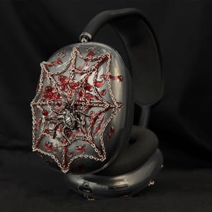 May include: Black headphones with a silver spider web design on one earcup. The web is decorated with red jewels and a silver spider.
