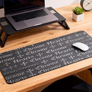 Puede incluir: Una alfombrilla de ratón negra con el texto "Chrome Hearts" y diseños de cruces. La alfombrilla está sobre un escritorio de madera con un portátil, una taza de café, bolígrafos y un bloc de notas. Un ratón de ordenador está sobre la alfombrilla.