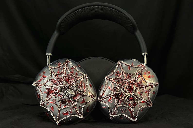 May include: Over-ear headphones with a black headband and silver earcups decorated with a red spiderweb design and silver spiders.