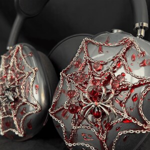 May include: A pair of black headphones with a spider web design. The headphones are decorated with silver chains and red accents. The spider web design is detailed and intricate.