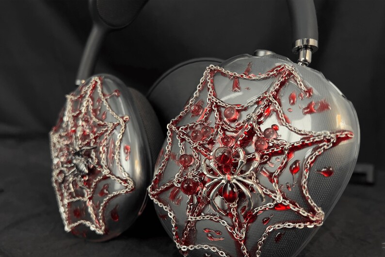 May include: A pair of black headphones with a spider web design. The headphones are decorated with silver chains and red accents. The spider web design is detailed and intricate.