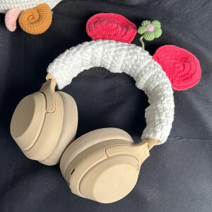 May include: Beige wireless headphones with a white crocheted headband and decorative crocheted elements. The decorations include red, pink, and brown crocheted shapes, and a small green flower. The headphones are on a black surface.