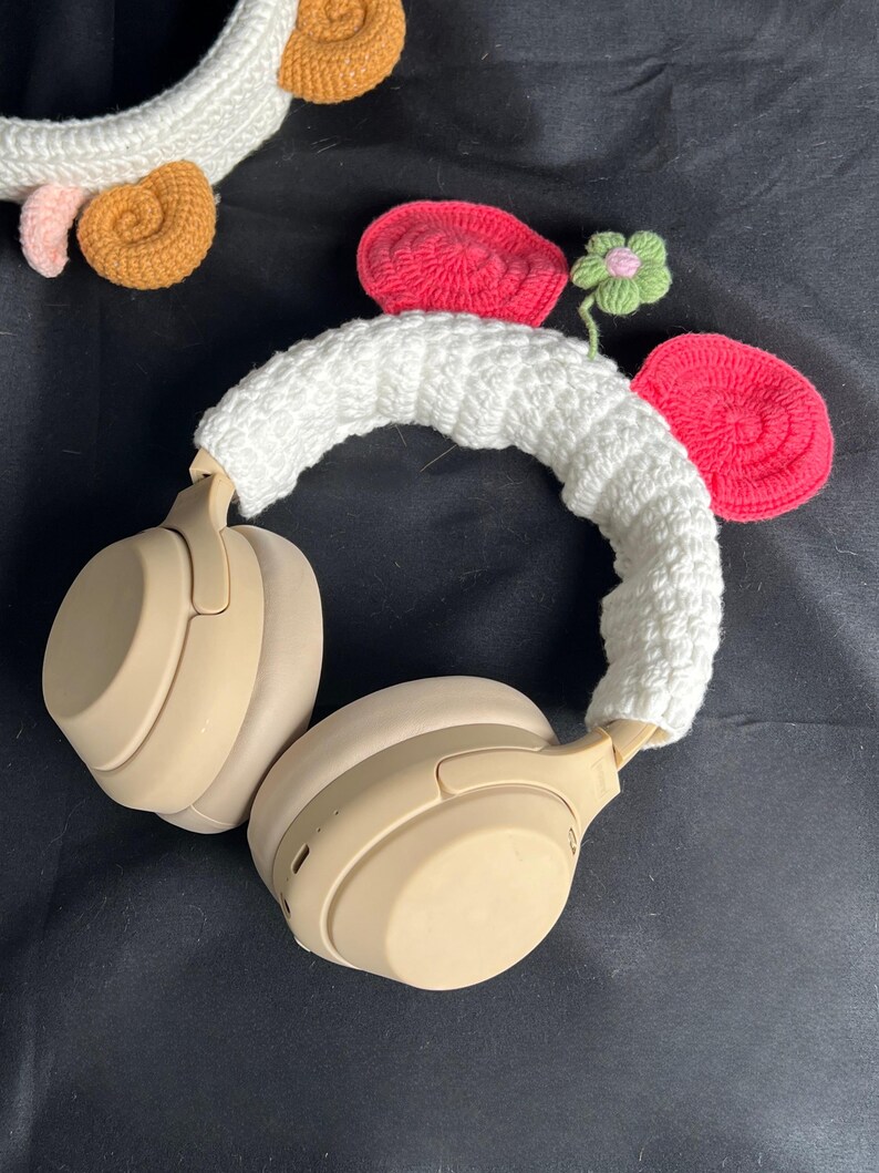 May include: Beige wireless headphones with a white crocheted headband and decorative crocheted elements. The decorations include red, pink, and brown crocheted shapes, and a small green flower. The headphones are on a black surface.