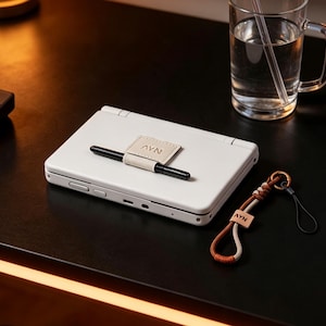 Op de afbeelding: Een witte draagbare spelcomputer met een zwarte stylus vastgehouden door een beige leren lus met de tekst AYN. Daarnaast ligt een gevlochten bruin en beige koord met een klein beige leren label met eveneens de tekst AYN.