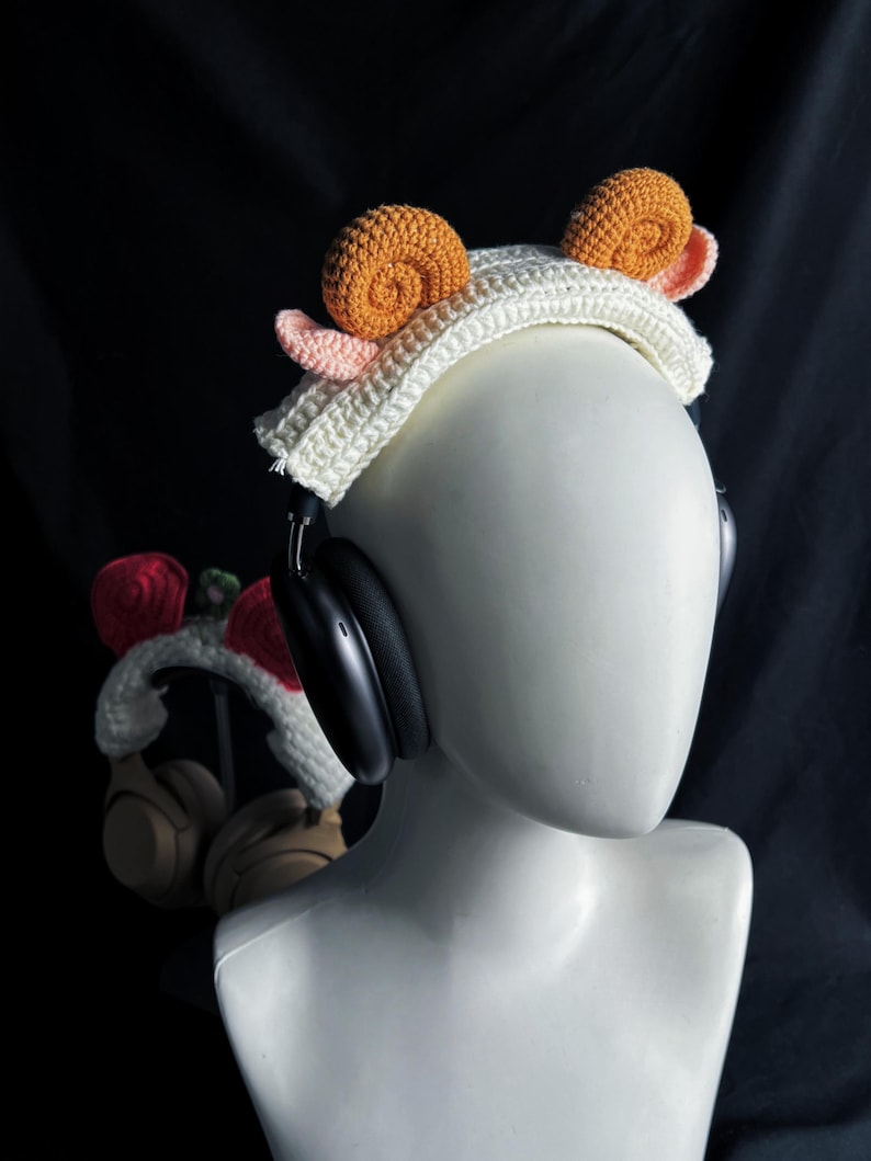 May include: A white crochet headband with two orange snail shell designs and pink accents. The headband is worn on a mannequin head with black headphones. A second pair of headphones with a similar design is visible in the background.