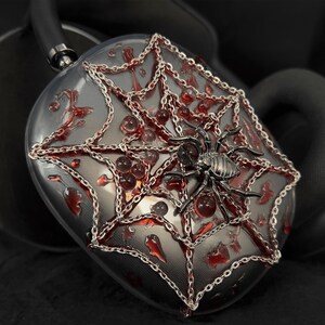 May include: A pair of black headphones with a spider web design. The headphones are decorated with silver chains and red accents. The spider web design features a silver spider in the center.