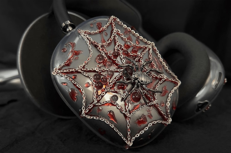 May include: A pair of black headphones with a spider web design. The headphones are decorated with silver chains and red accents. The spider web design features a silver spider in the center.