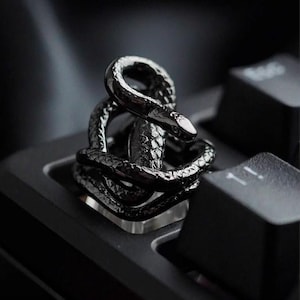 May include: A coiled black snake ring with a textured surface and a white tip on the head. The ring is set against a dark background, resting on a keyboard key.