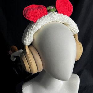 May include: Headphones with a white crocheted headband featuring red ear accents and a small green and pink flower. The headphones are tan and sit on a white mannequin head against a black background.