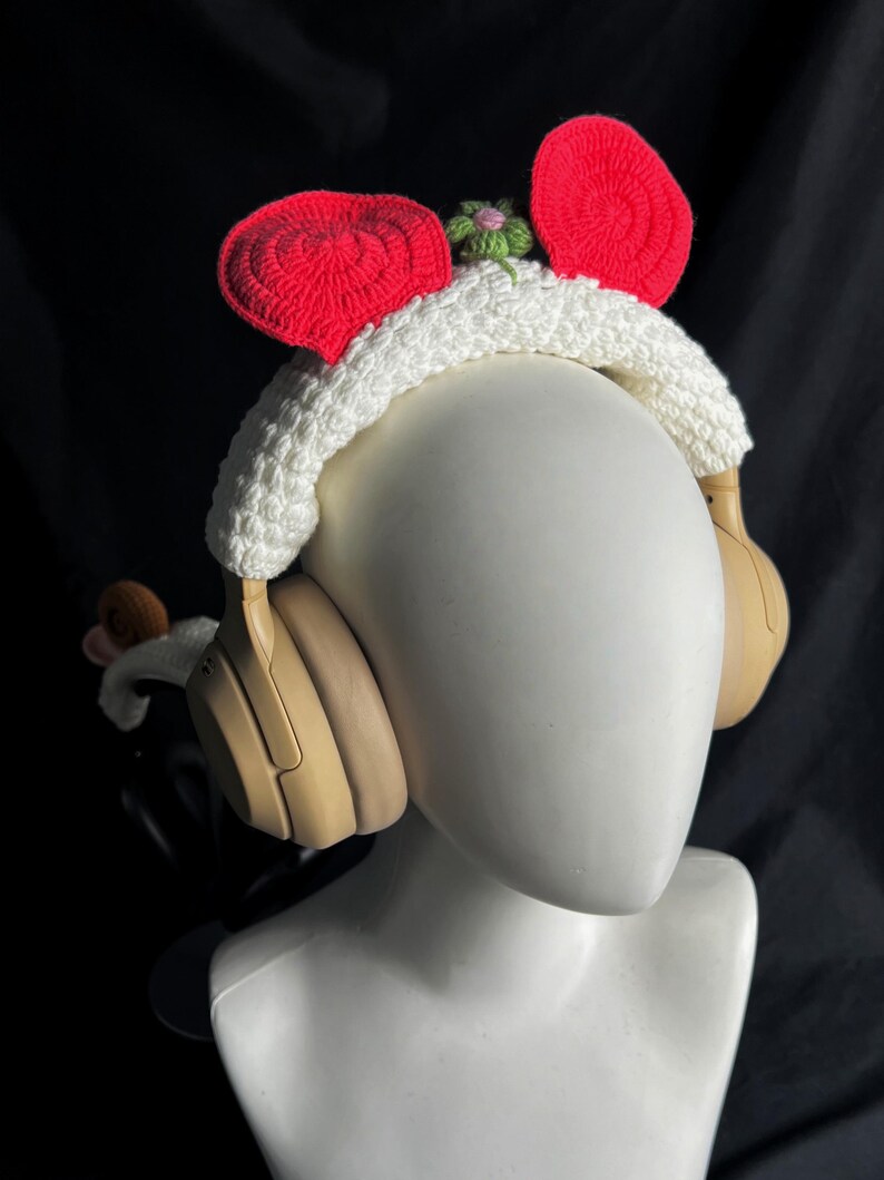 May include: Headphones with a white crocheted headband featuring red ear accents and a small green and pink flower. The headphones are tan and sit on a white mannequin head against a black background.