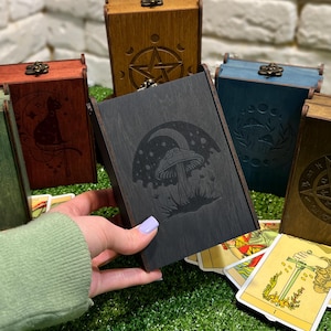 Könnte beinhalten: Eine Sammlung von Holzkisten in verschiedenen Farben, darunter Grün, Rot, Blau und Braun, jede mit einem einzigartigen eingravierten Design. Die rechteckigen Kisten haben Metallverschlüsse. Tarotkarten sind vor den Kisten verstreut.