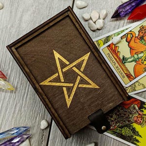 May include: A wooden box with a gold pentagram design on the lid. The box is surrounded by tarot cards, including the Knight of Wands and the Empress.