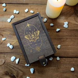 May include: A wooden box with a gold butterfly design and a star on the lid. The box is on a wooden surface with white stones scattered around it.