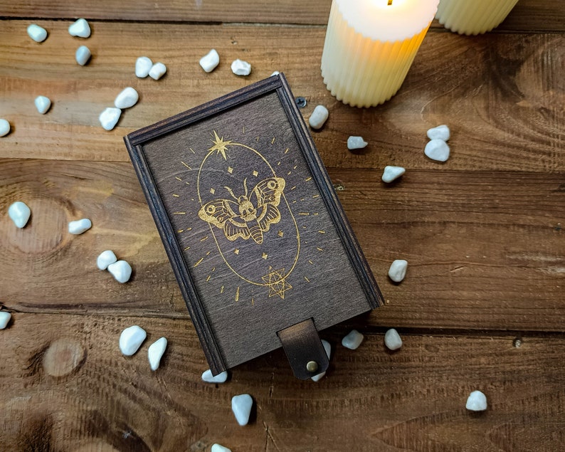 May include: A wooden box with a gold butterfly design and a star on the lid. The box is on a wooden surface with white stones scattered around it.