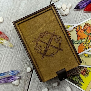 May include: A wooden box with a hinged lid, featuring a laser-engraved design of two crossed swords with a pentagram and a circle. The box is closed and sitting on a table with tarot cards spread out around it.