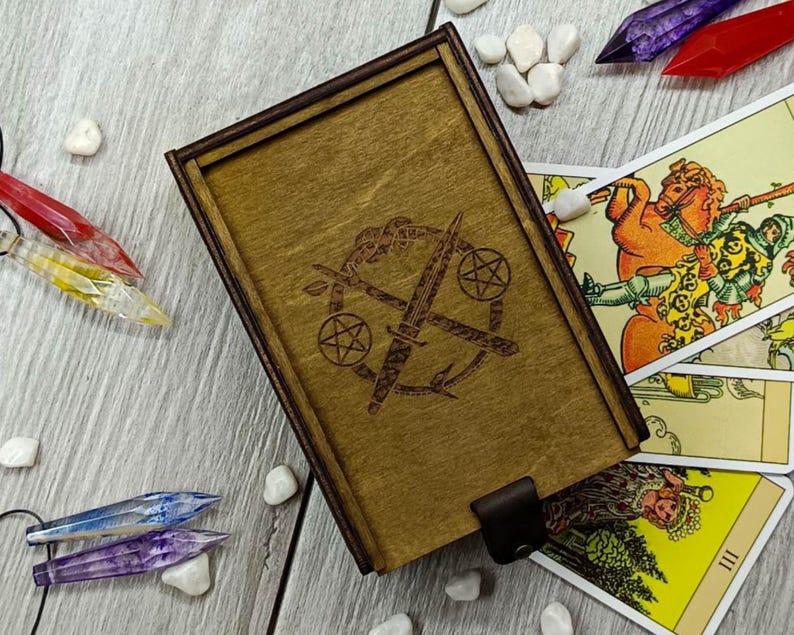 May include: A wooden box with a hinged lid, featuring a laser-engraved design of two crossed swords with a pentagram and a circle. The box is closed and sitting on a table with tarot cards spread out around it.