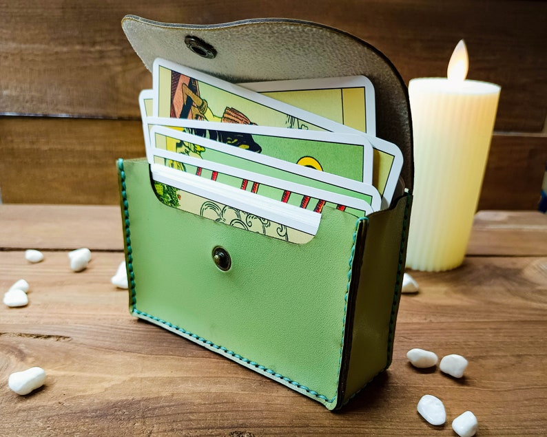 May include: A light green leather tarot card case with a flap secured by a snap. The case holds a deck of tarot cards with colorful illustrations. A lit candle and small white stones are in the background.