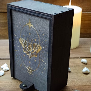 May include: A dark gray wooden box with a gold-colored butterfly design on the front. The box has a leather strap with a metal button.