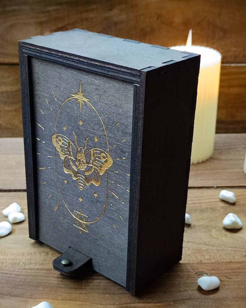 May include: A dark gray wooden box with a gold-colored butterfly design on the front. The box has a leather strap with a metal button.