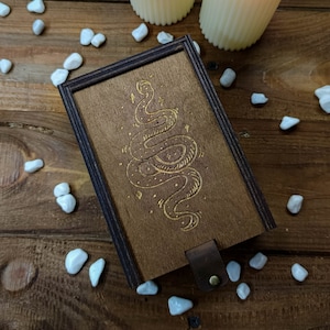 May include: A wooden box with a dark brown finish and a gold snake design engraved on the lid. The box has a leather strap closure.