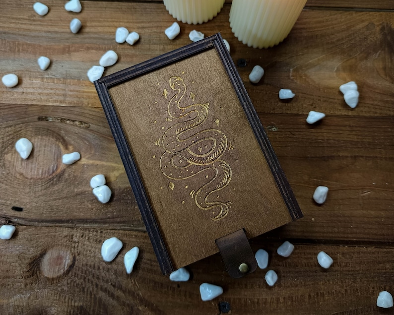 May include: A wooden box with a dark brown finish and a gold snake design engraved on the lid. The box has a leather strap closure.