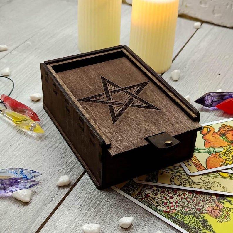 May include: A wooden box with a pentagram design on the lid. The box is closed and has a leather strap. The box is sitting on a table with tarot cards and other objects.