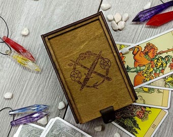 May include: A wooden box with a leather strap closure. The box has a laser-engraved design of two crossed swords with a pentagram on each sword. The box is sitting on a table with tarot cards.