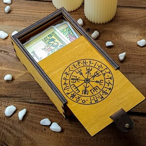 May include: A wooden box with a yellow lid and a compass design. The lid is open and there are playing cards inside. The box is on a wooden surface.