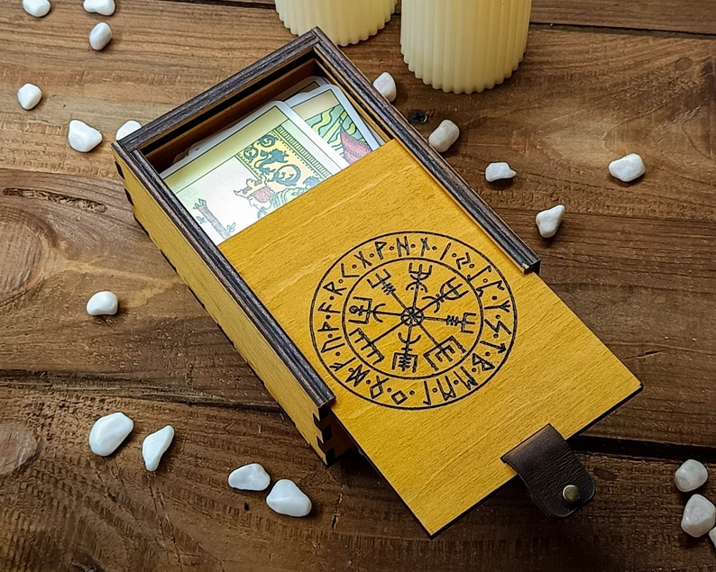 May include: A wooden box with a yellow lid and a compass design. The lid is open and there are playing cards inside. The box is on a wooden surface.