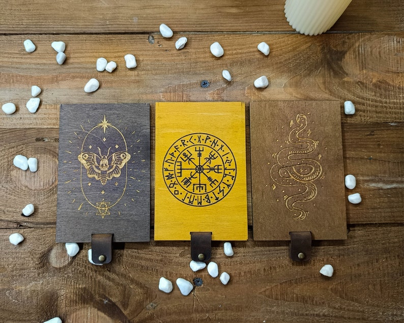 May include: Three wooden plaques with different designs. The first plaque has a butterfly with a skull in the center. The second plaque has a compass with the text "・N・P・F・R・<・X・P-N-X-". The third plaque has a snake with a tail that forms a circle.