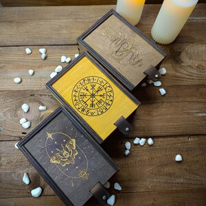 May include: Three wooden boxes with different designs. The first box has a compass with runes around it, the second box has a snake design, and the third box has a butterfly design. The boxes are on a wooden surface with white stones.