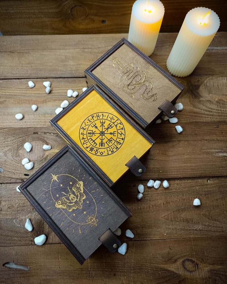May include: Three wooden boxes with different designs. The first box has a compass with runes around it, the second box has a snake design, and the third box has a butterfly design. The boxes are on a wooden surface with white stones.