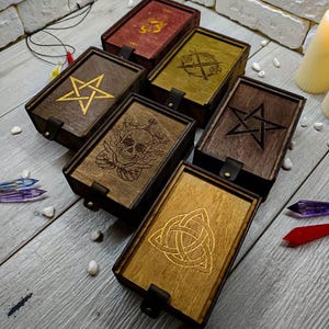 May include: Five wooden boxes with different engraved designs. The designs include a pentagram, a skull, a Celtic knot, a sword and a dagger, and a five-pointed star. The boxes are all made of dark wood with a light brown stain.