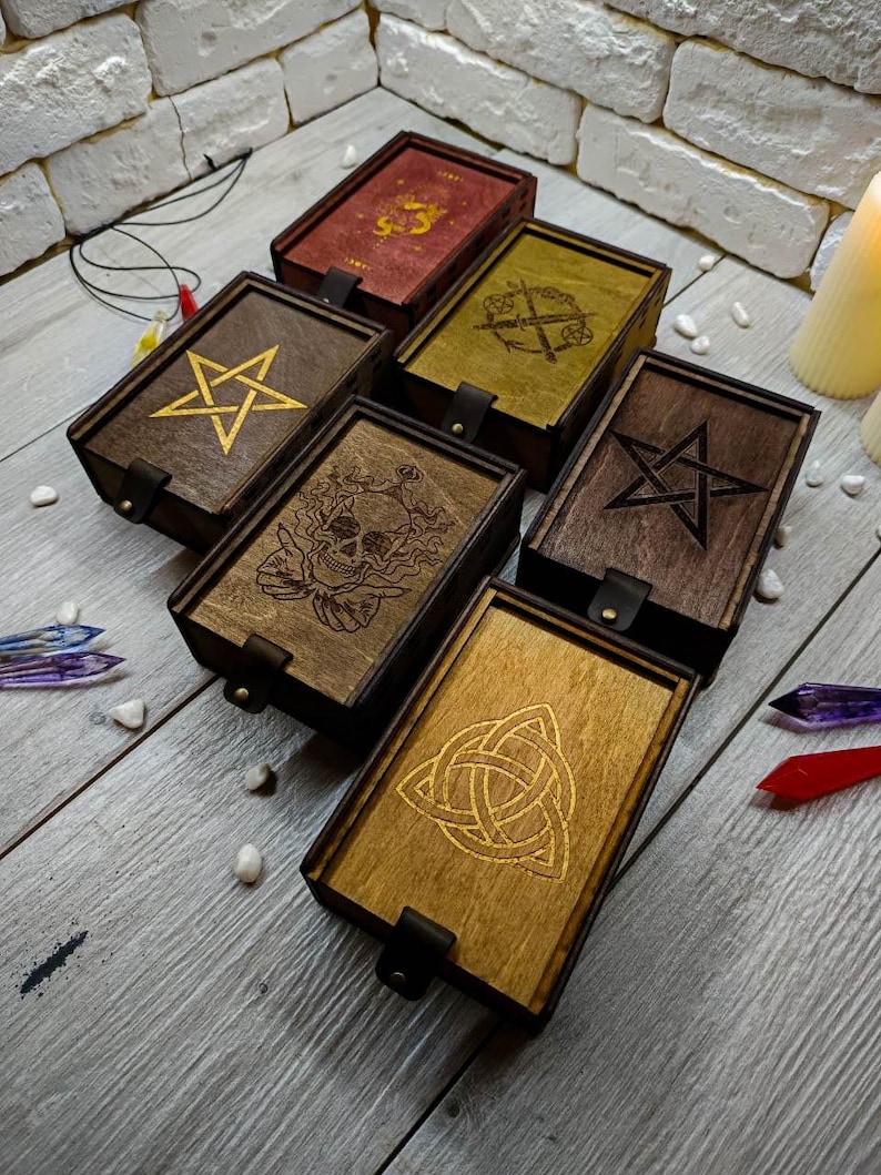 May include: Five wooden boxes with different engraved designs. The designs include a pentagram, a skull, a Celtic knot, a sword and a dagger, and a five-pointed star. The boxes are all made of dark wood with a light brown stain.