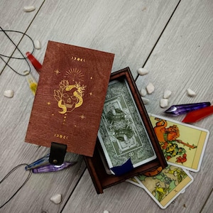 May include: A wooden box with a red lid and gold skull design. The box is open and contains a deck of tarot cards. The cards are spread out on a wooden surface.