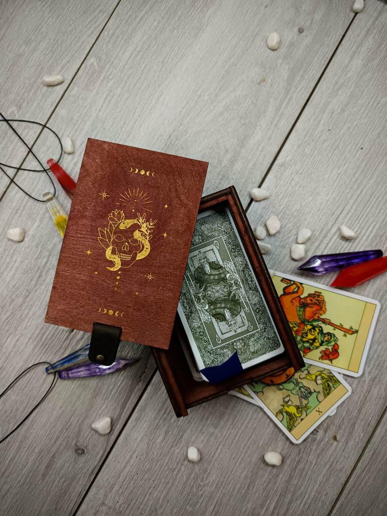 May include: A wooden box with a red lid and gold skull design. The box is open and contains a deck of tarot cards. The cards are spread out on a wooden surface.