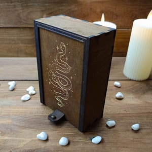 May include: A wooden box with a dark brown finish and a gold snake design on the front. The box has a leather strap with a metal buckle.