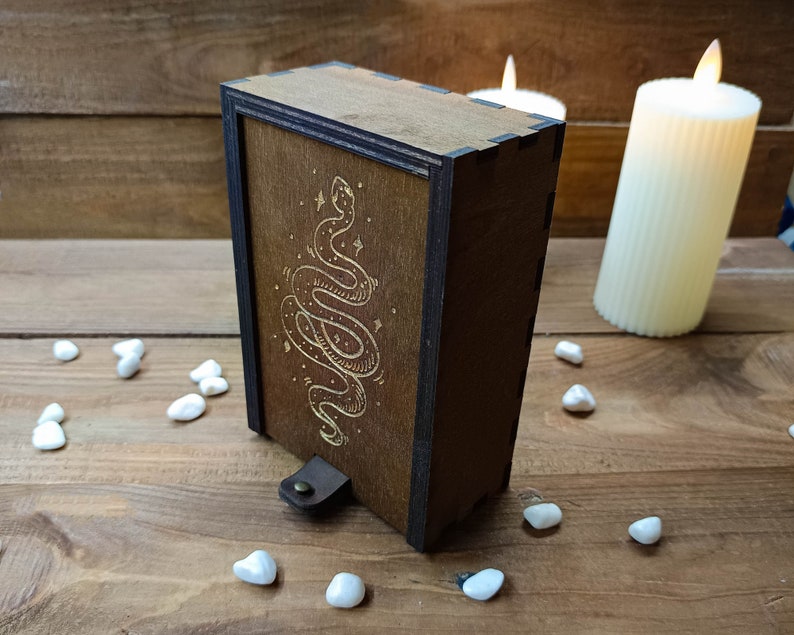 May include: A wooden box with a dark brown finish and a gold snake design on the front. The box has a leather strap with a metal buckle.