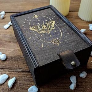 May include: A dark wooden box with a gold-colored design of a butterfly and stars. The box has a leather strap and a hinged lid.