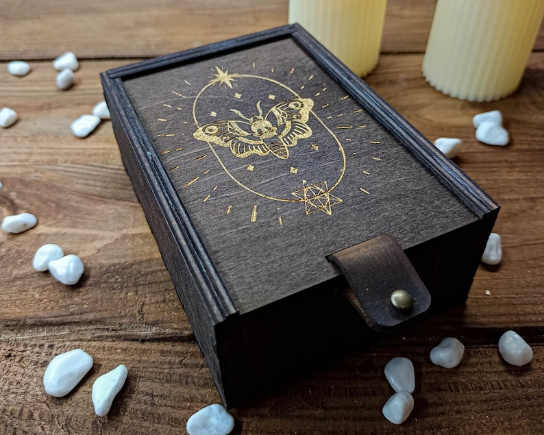 May include: A dark wooden box with a gold-colored design of a butterfly and stars. The box has a leather strap and a hinged lid.