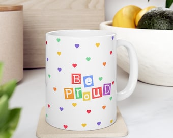 May include: White ceramic mug with a rainbow-colored "Be Proud" message in block letters and a white background with a scattered pattern of small multi-colored hearts.