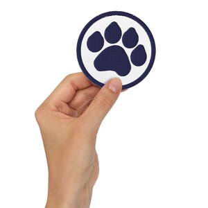 May include: A round patch featuring a navy blue paw print inside a white circle, with a navy blue border. The patch is held against a white background.