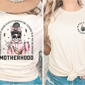 K&ouml;nnte beinhalten: Ein cremefarbenes T-Shirt mit einem Motiv eines Skeletts, das einen rosa Hoodie, eine Sonnenbrille und eine Bauchtasche tr&auml;gt. Der Text "Some days I rock it. Some days it rocks me. Either way we're rockin' Motherhood" ist auf der R&uuml;ckseite des Shirts aufgedruckt. Die Vorderseite des Shirts zeigt ein Motiv einer Skeletthand mit dem aufgedruckten Text "Rockin' Motherhood".