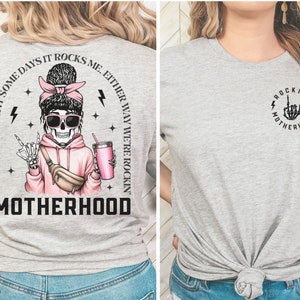 K&ouml;nnte beinhalten: Graues T-Shirt mit einem Skelett, das einen rosa Hoodie und eine Sonnenbrille tr&auml;gt. Der Text auf dem Shirt lautet "Some days I rock it some days it rocks me, either way we're rockin' motherhood". Die R&uuml;ckseite des Shirts hat eine Skeletthand mit einer Rock'n'Roll-Handgeste. Der Text auf der R&uuml;ckseite des Shirts lautet "Rockin' Motherhood".