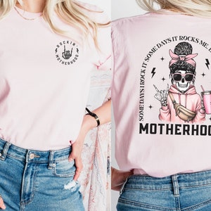 K&ouml;nnte beinhalten: Pinkes T-Shirt mit einer schwarzen Grafik eines Skeletts, das einen rosa Hoodie tr&auml;gt und ein Getr&auml;nk h&auml;lt. Der Text auf dem Shirt lautet "Some days I rock it some days it rocks me. Either way we're rockin' motherhood."
