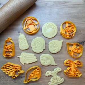 Könnte beinhalten: Ein Set mit 12 gelben und orangefarbenen Plastik-Ausstechern in Form von Figuren aus dem Film Inside Out. Die Ausstecher sind in einem Kreis auf einer Holzoberfläche angeordnet.