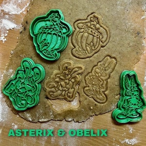 Könnte beinhalten: Grüne Plastik-Ausstecher in Form von Asterix- und Obelix-Charakteren aus den Asterix-Comics. Die Ausstecher befinden sich auf einer braunen Oberfläche mit dem darunter gedruckten Text "Asterix & Obelix".